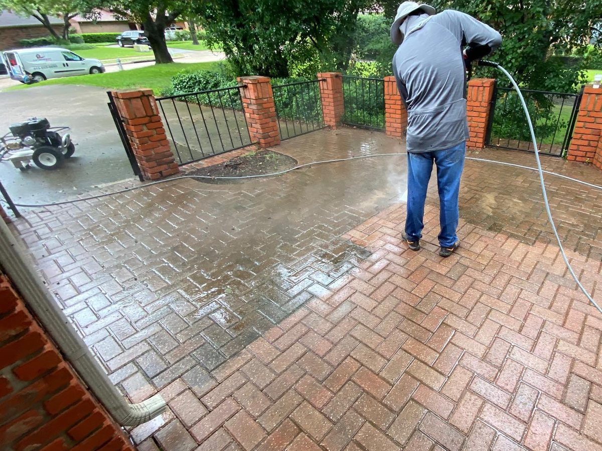 Worker pressure washing a brick paver patio, clean stripe showing against the dirty surface